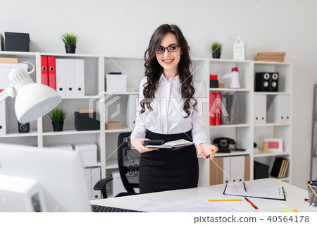 A beautiful young girl stands near an office desk and holds a pen, notebook and calculator in her A beautiful young girl stands near an office desk and holds a pen, notebook and calculator in her 40564178