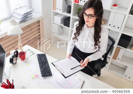 A beautiful young girl stands near an office desk and holds in her hands a sheet for notes and a A beautiful young girl stands near an office desk and holds in her hands a sheet for notes and a 40566369