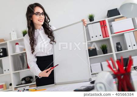 Beautiful young girl stands near an office desk and points with a pen on an empty board. 40566384