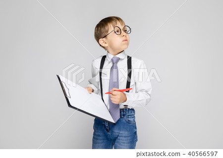 A charming boy in a white shirt, suspenders, a tie and light jeans stands on a gray background. The 40566597