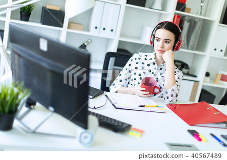 A young girl sits in headphones at a table in the office, holds a red cup in her hands and looks at 40567489