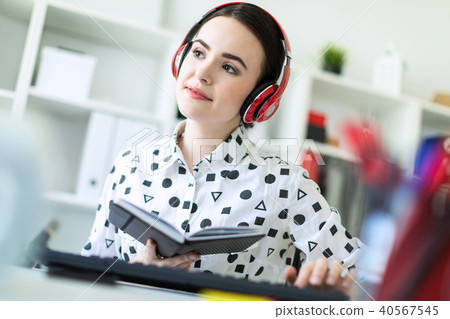 Beautiful young girl sits in headphones at desk in office, holds a notebook in her hand and prints 40567545