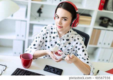 Beautiful young girl sitting in headphones at desk in office eating yogurt with red filling. 40567556