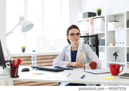 A beautiful young girl in a white robe is sitting at a table and holding a wooden figure of a man in 40567559