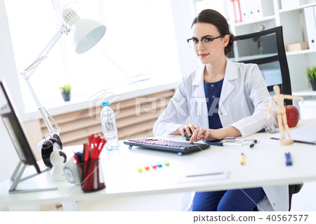 A beautiful young girl in a white robe is sitting at the table and typing on the keyboard. 40567577