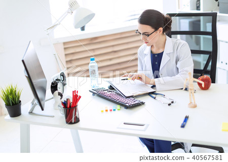 A beautiful young girl in a white robe is sitting at a computer desk with documents and a pen in her 40567581