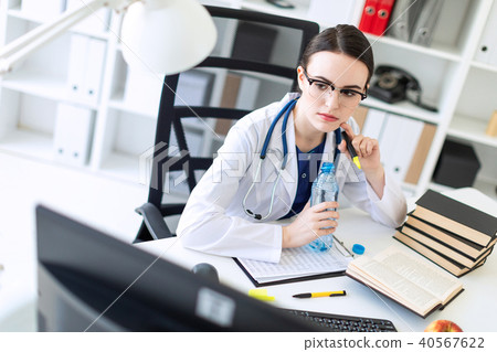 A beautiful young girl in a white robe is sitting at the computer desk, holding an open bottle of 40567622