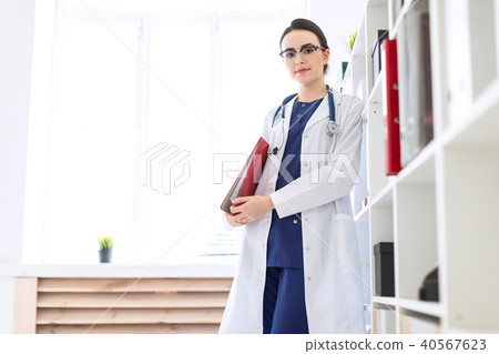 A beautiful young girl in a white coat is standing near the shelter and holding a red folder under 40567623