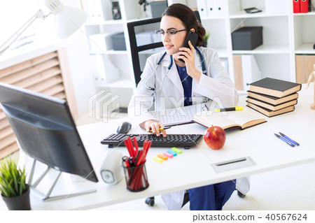 A beautiful young girl in a white robe is sitting at the table, talking on the phone and holding her 40567624