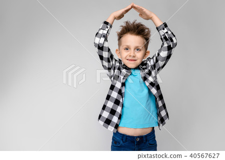 A handsome boy in a plaid shirt, blue shirt and jeans stands on a gray background. The boy folded 40567627