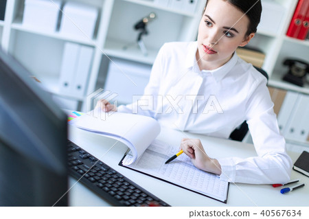A young girl in the office holds a pen in her hand and works with documents and a computer. 40567634