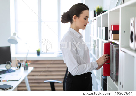 A young girl in the office is standing near the shelf and pulling out a red folder. 40567644