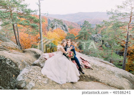 Horizontal view of the happy newlywed couple sitting on the knitted plaid on the rock. Horizontal view of the happy newlywed couple sitting on the knitted plaid on the rock. 40586241