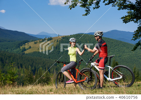 Happy bikers couple, man and woman standing with bikes on grassy hill under big green tree branch 40610756