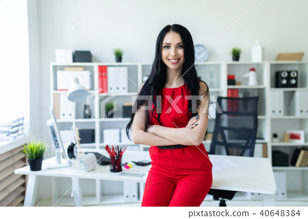 Beautiful young girl in a red suit is standing in the office, leaning on the table. Beautiful young girl in a red suit is standing in the office, leaning on the table. 40648384