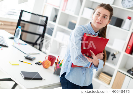 A young girl in the office is standing, leaning on a table, and is holding a phone and a folder with 40648475