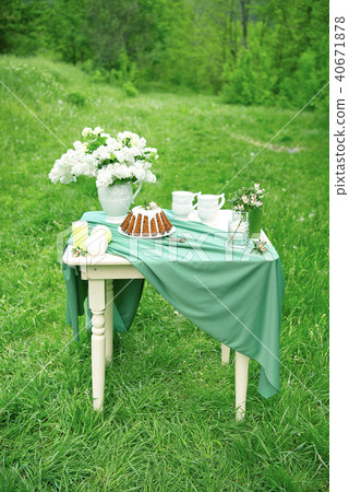 Cake with some cups on picnic table decorated with flowers, candles, vase in green field 40671878