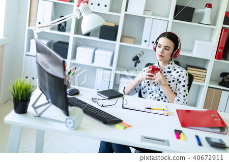 A young girl sits in headphones at a table in the office, holds a red cup in her hands and looks at 40672911