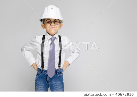 A charming boy in a white shirt, suspenders, a tie and light jeans stands on a gray background. Boy 40672936