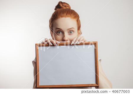 Portrait of cheerful redhead girl with hair bun holding empty wooden frame. 40677003