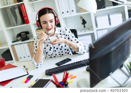 Beautiful young girl sitting in headphones at desk in office, eating yogurt and looking at monitor. 40703305