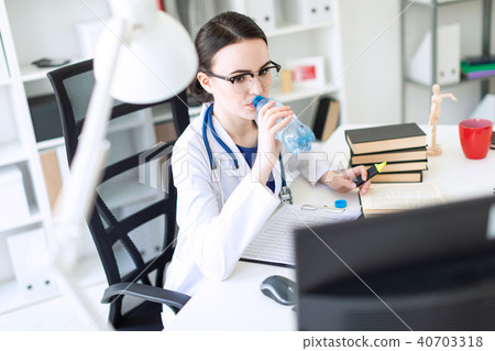 A beautiful young girl in a white robe sits at a computer desk, drinks water and looks at the 40703318