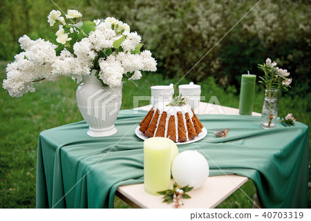 Cake with some cups on picnic table decorated with flowers, candles, vase in green field 40703319