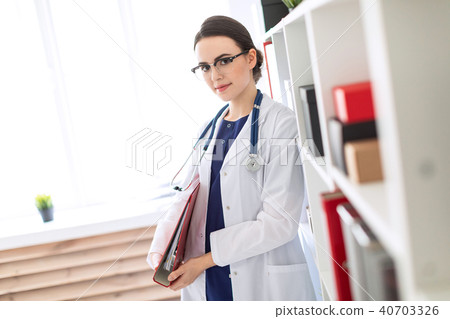A beautiful young girl in a white coat is standing near the shelter and holding a red folder under 40703326