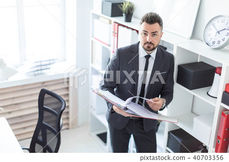 The man is standing near the rack in the office and scrolls through the folder with the documents. The man is standing near the rack in the office and scrolls through the folder with the documents. 40703356
