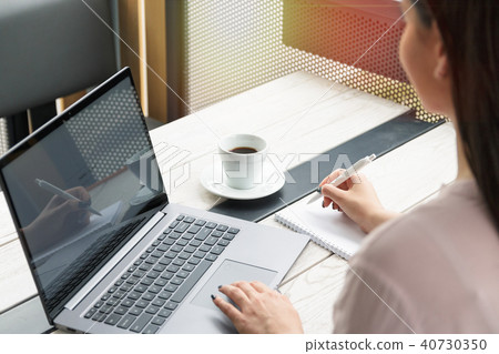 Close up portrait of a young woman working on laptop and writing, coffee on the table. 40730350