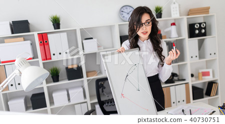 A beautiful young girl stands near an office desk and draws a magnetic marker on the magnetic board. A beautiful young girl stands near an office desk and draws a magnetic marker on the magnetic board. 40730751