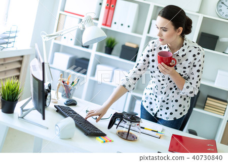 A young girl is standing in the office near the table, holding a red mug in her hand and typing on 40730840
