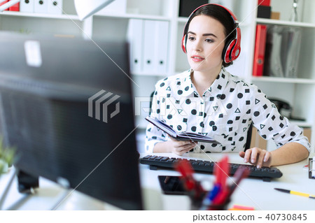 Beautiful young girl sits in headphones at desk in office, holds a notebook in her hand and prints 40730845