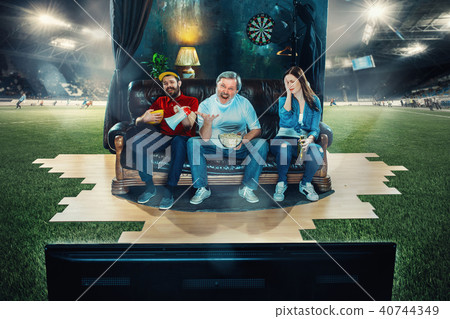 Soccer football fans sitting on the sofa and watching TV in the middle of a football field. Soccer football fans sitting on the sofa and watching TV in the middle of a football field. 40744349
