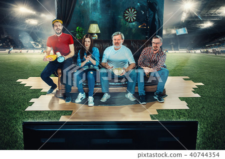 Soccer football fans sitting on the sofa and watching TV in the middle of a football field. Soccer football fans sitting on the sofa and watching TV in the middle of a football field. 40744354