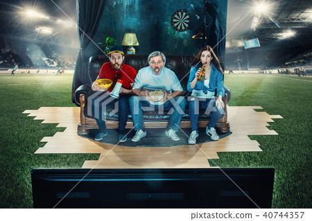 Soccer football fans sitting on the sofa and watching TV in the middle of a football field. Soccer football fans sitting on the sofa and watching TV in the middle of a football field. 40744357