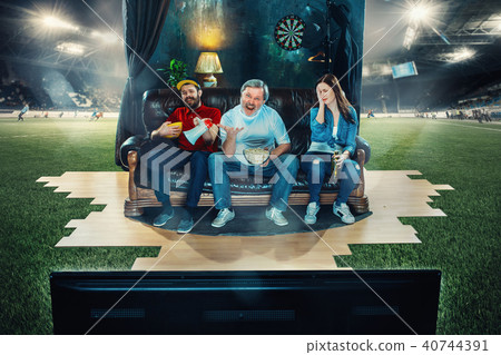 Soccer football fans sitting on the sofa and watching TV in the middle of a football field. Soccer football fans sitting on the sofa and watching TV in the middle of a football field. 40744391