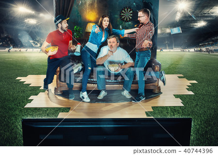 Soccer football fans sitting on the sofa and watching TV in the middle of a football field. 40744396
