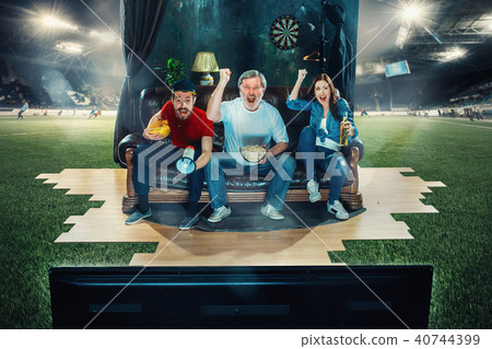Soccer football fans sitting on the sofa and watching TV in the middle of a football field. Soccer football fans sitting on the sofa and watching TV in the middle of a football field. 40744399