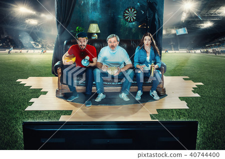 Soccer football fans sitting on the sofa and watching TV in the middle of a football field. Soccer football fans sitting on the sofa and watching TV in the middle of a football field. 40744400
