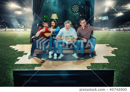 Soccer football fans sitting on the sofa and watching TV in the middle of a football field. Soccer football fans sitting on the sofa and watching TV in the middle of a football field. 40748900