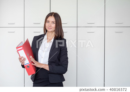 Beautiful business woman stands with a red folder in hands in a office Beautiful business woman stands with a red folder in hands in a office 40816372
