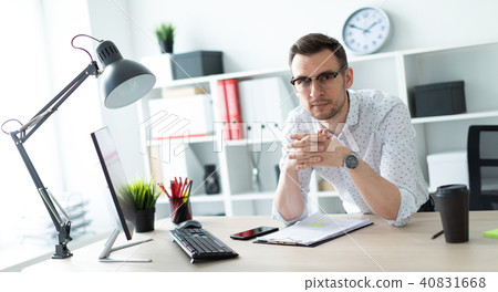 A young man in glasses stands near a table in the office. A young man in glasses stands near a table in the office. 40831668