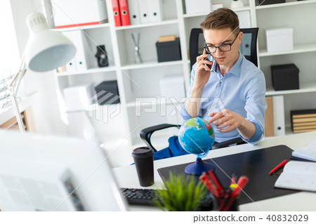 A young man sits in the office at a computer desk, talking on the phone and looking at the globe. A young man sits in the office at a computer desk, talking on the phone and looking at the globe. 40832029