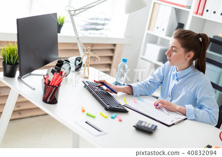 A young girl in the office sits at a table, holds a pen in her hand and works at the computer. 40832094