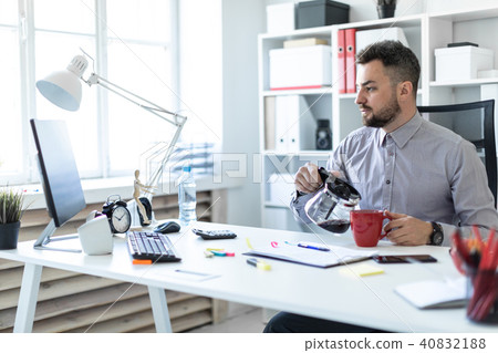 A young man in the office sits at a table, looks at the monitor and pours coffee into a cup. A young man in the office sits at a table, looks at the monitor and pours coffee into a cup. 40832188