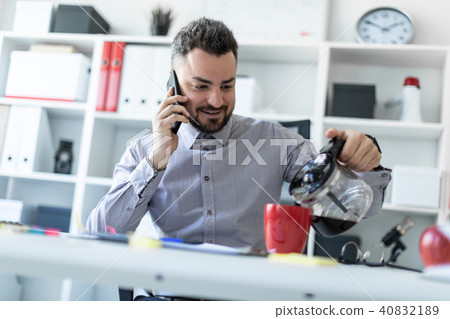 A young man in the office sits at a table, talking on the phone and pouring coffee into a cup. A young man in the office sits at a table, talking on the phone and pouring coffee into a cup. 40832189