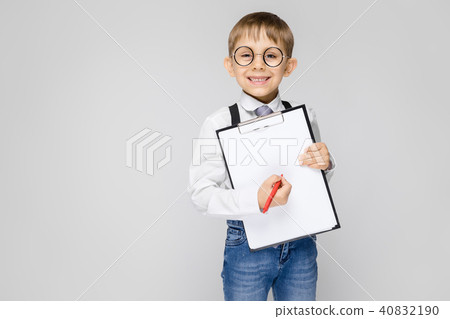 A charming boy in a white shirt, suspenders, a tie and light jeans stands on a gray background. The A charming boy in a white shirt, suspenders, a tie and light jeans stands on a gray background. The 40832190