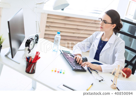 A beautiful young girl in a white robe is sitting at the table and typing on the keyboard. 40850094