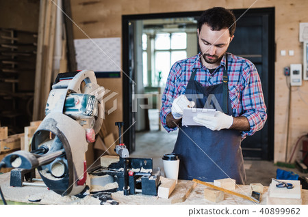 carpenter texting someone on his smart phone near circular saw in a dusty workshop carpenter texting someone on his smart phone near circular saw in a dusty workshop 40899962